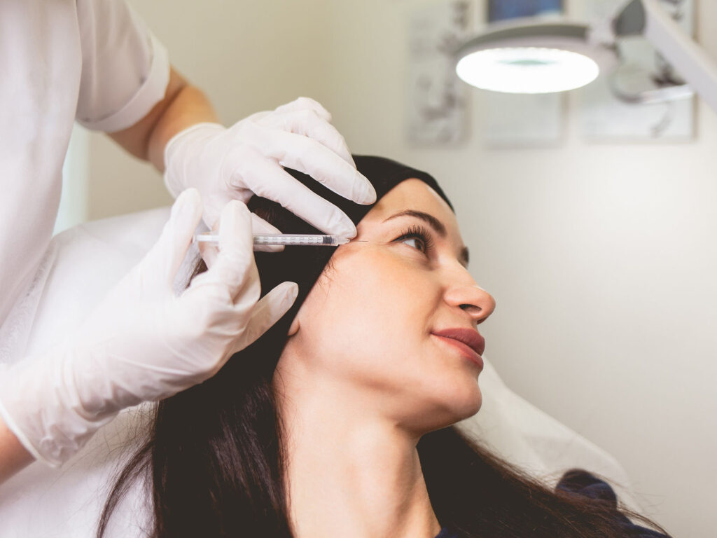 Clinician wearing gloves administering a Botox injection near the temple/forehead in a medical clinic setting.